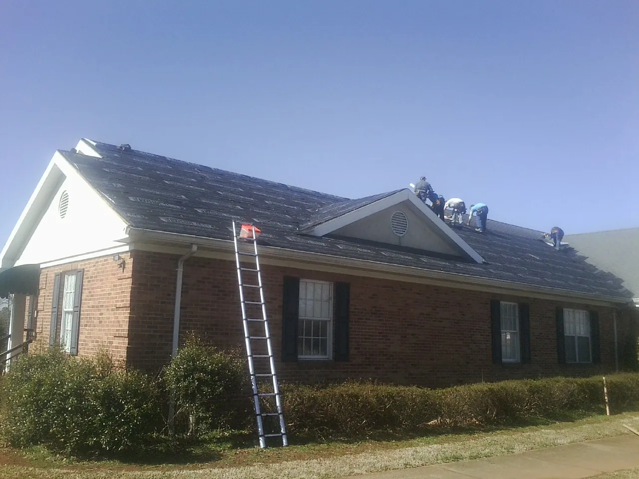 Crew tearing shingles off a brick ranch roof with a ladder set — Emergency Roof Repair work by our crew in Ballston Lake