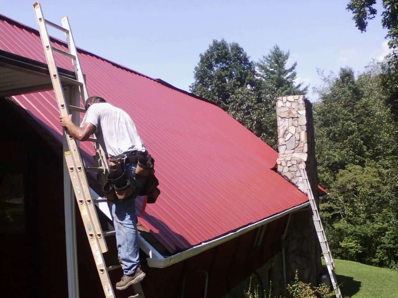 Roofer on a ladder beside a red metal roof and stone chimney — Soffit & Fascia Repair by Roofing Brothers in Ballston Lake